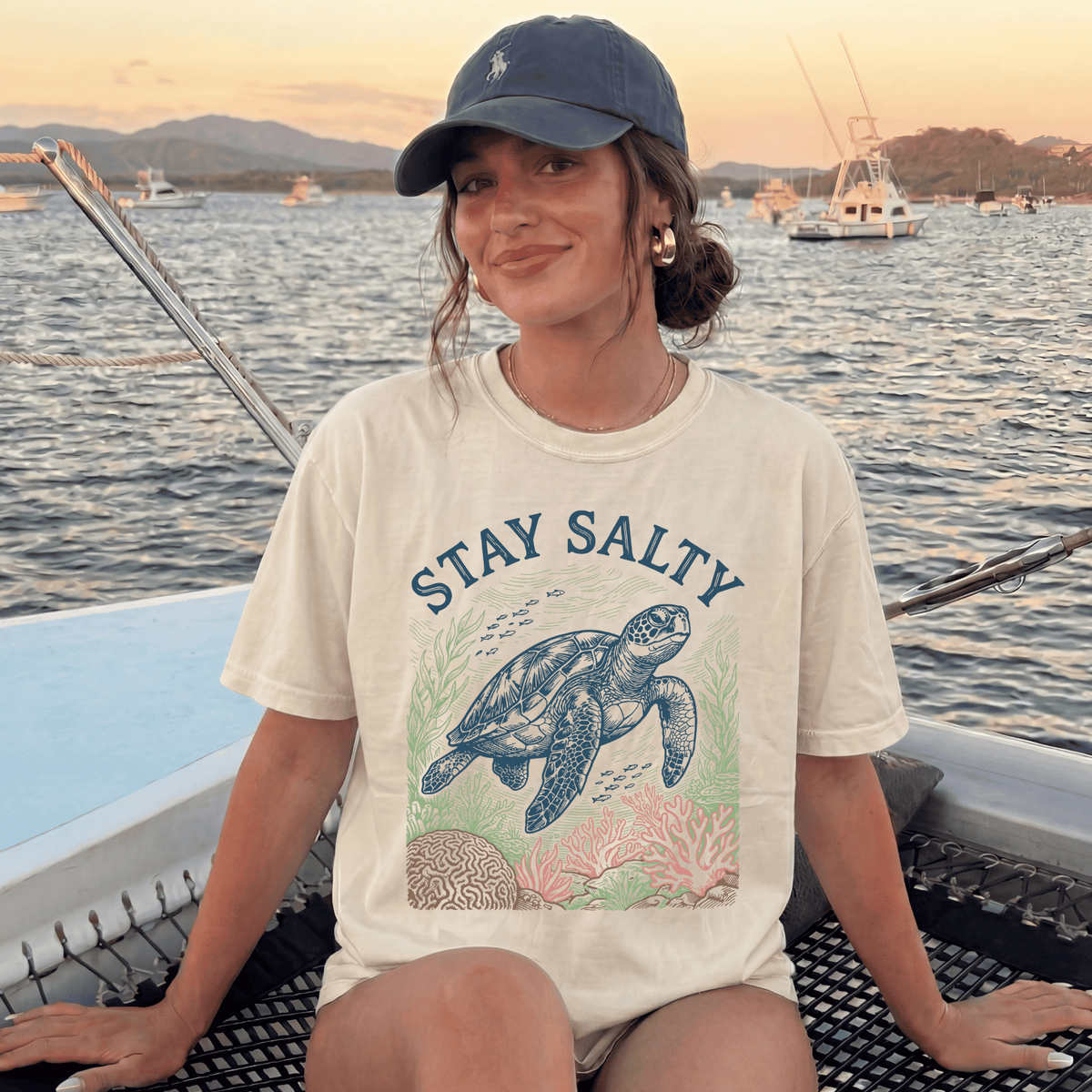 A woman wearing a white t-shirt with a graphic of a sea turtle and the text "STAY SALTY" is sitting on a boat, smiling at the camera.