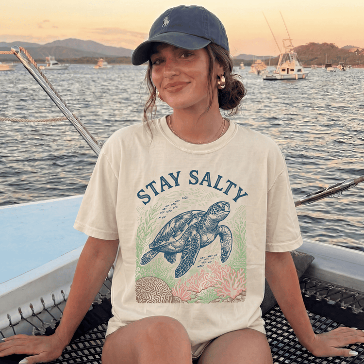 A woman wearing a white t-shirt with a graphic of a sea turtle and the text "STAY SALTY" is sitting on a boat, smiling at the camera.