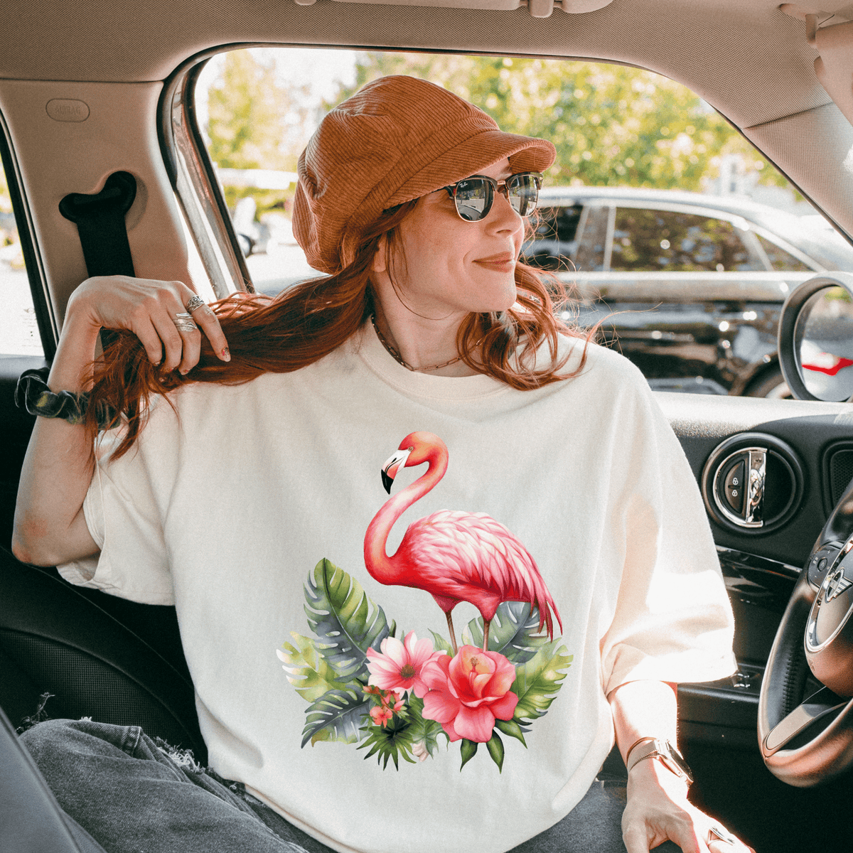 A woman wearing a white t-shirt with a pink flamingo and tropical flowers is sitting in a car, smiling and looking out the window.