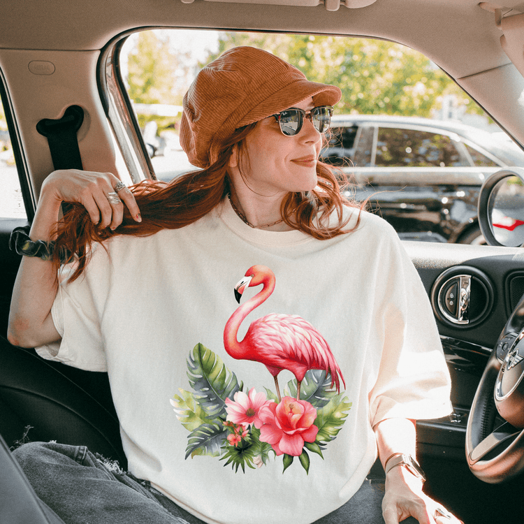 A woman wearing a white t-shirt with a pink flamingo and tropical flowers is sitting in a car, smiling and looking out the window.