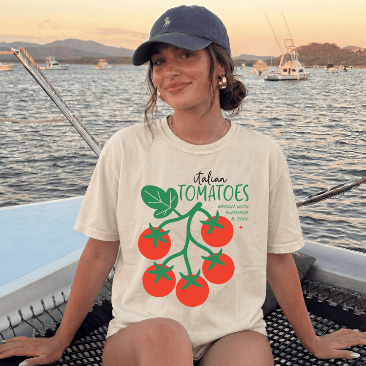 A woman wearing a white t-shirt with a graphic of tomatoes and text, sitting on a boat in the water with mountains in the background.