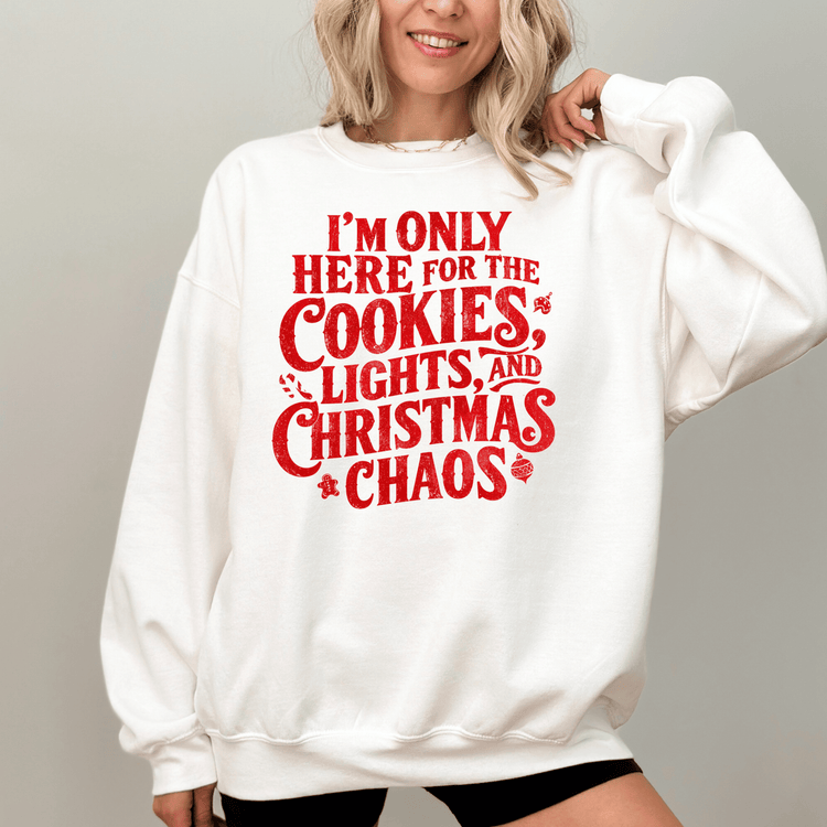 A woman wearing a white sweatshirt with a red text design that says "I'm only here for the cookies, lights, and Christmas chaos".