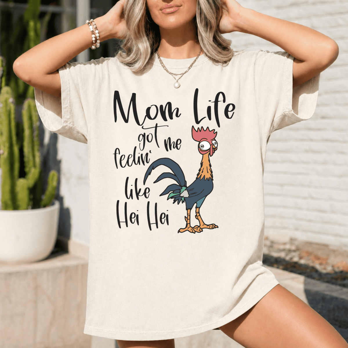A woman wearing a white t-shirt with a graphic design of a rooster and the text "Mom Life got me feelin like Hei Hei" printed on it.