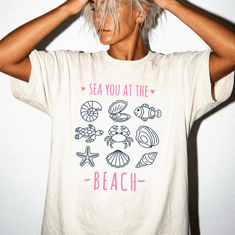 A woman wearing a white t-shirt with a graphic design of various sea creatures and the text "SEA YOU AT THE BEACH" printed on it.