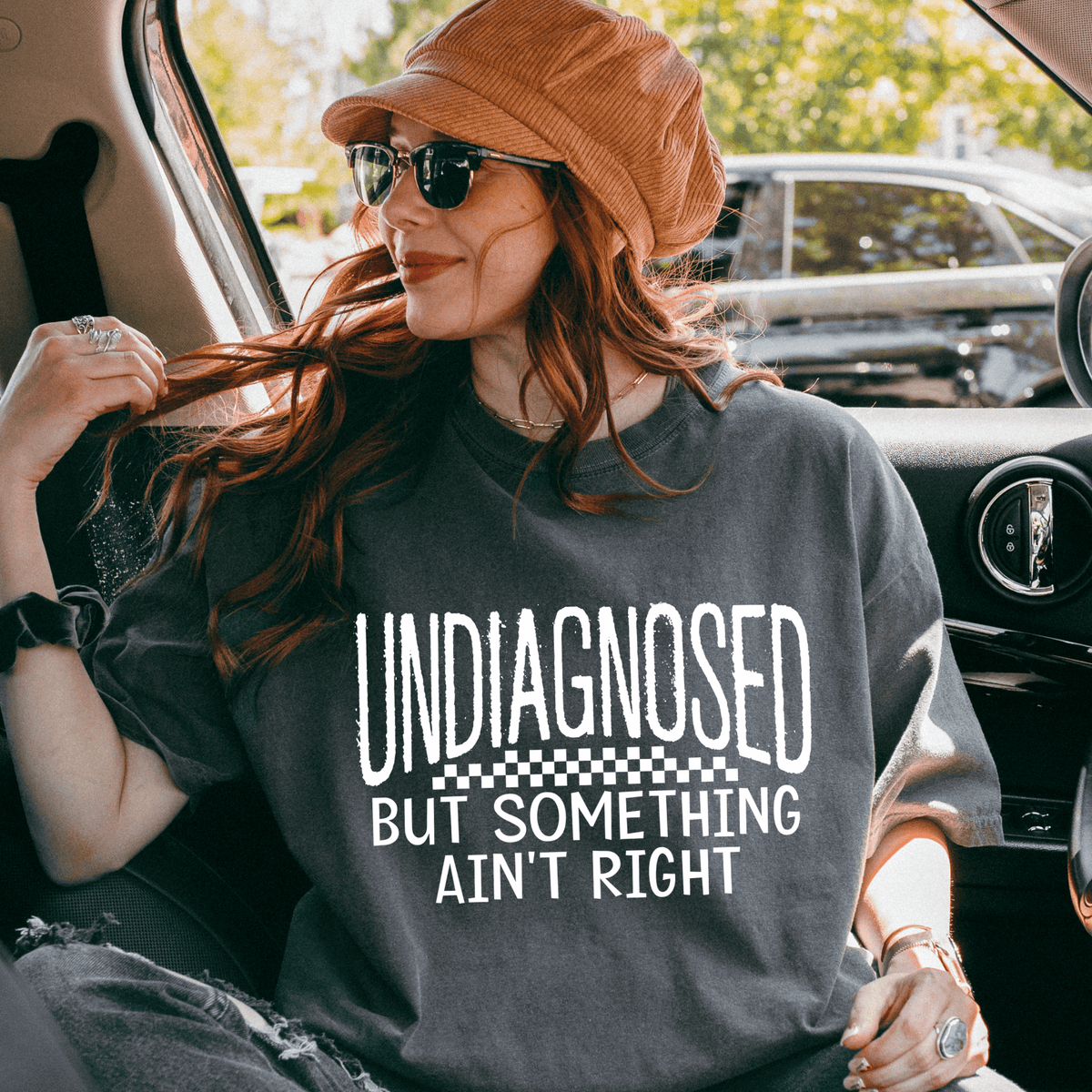 A woman wearing a hat and sunglasses is sitting in the back seat of a car, looking out the window. She is wearing a gray t-shirt with the text "UNDIAGNOSED BUT SOMETHING AIN'T RIGHT" printed on it.