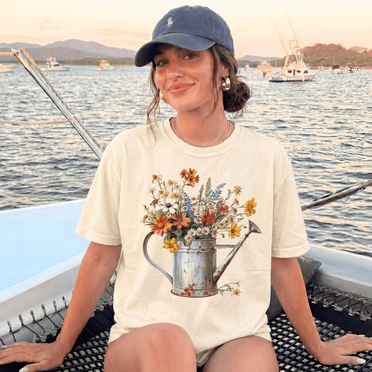 A woman wearing a white t-shirt with a flower design and a blue cap is sitting on a boat, smiling at the camera.