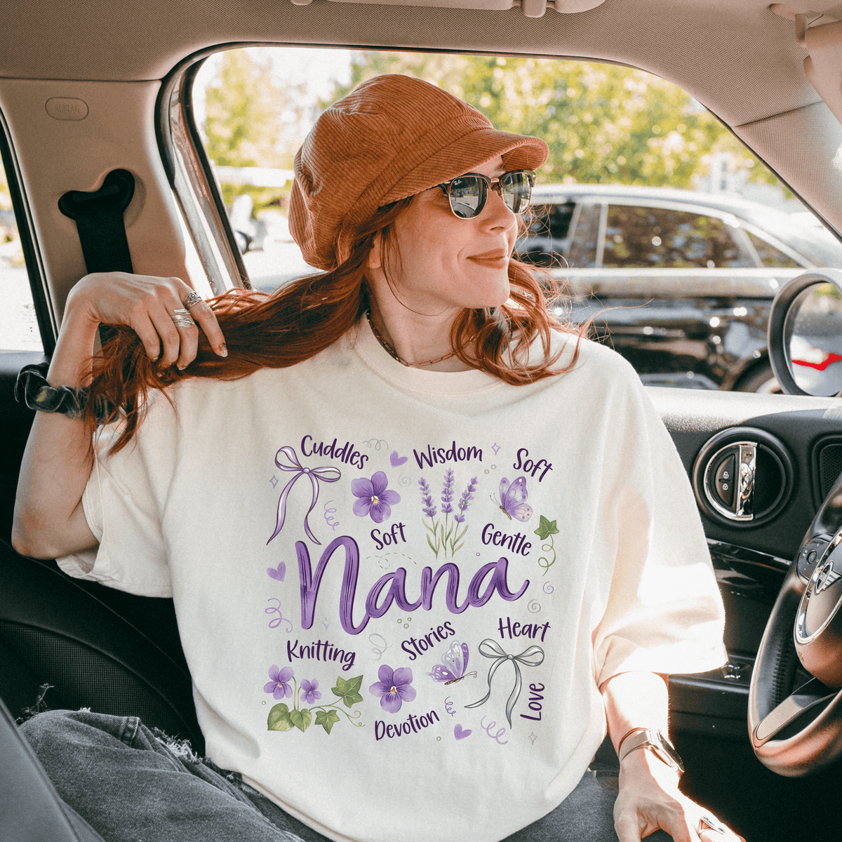 A woman wearing a white t-shirt with purple text and a brown hat is sitting in a car, smiling and looking out the window.