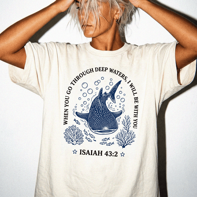 A person wearing a white t-shirt with a graphic design featuring a blue whale surrounded by bubbles and marine life, along with the text "When you go through deep waters, I will be with you" and the Bible verse Isaiah 43:2.