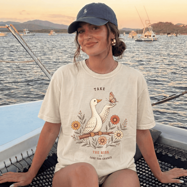 A woman wearing a white t-shirt with a graphic design of a bird and flowers, sitting on a boat with a scenic background of mountains and boats.