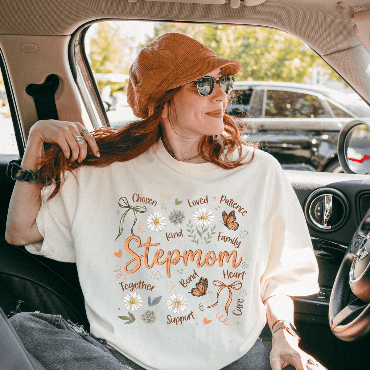 A woman wearing a white t-shirt with a design and text, sitting in a car with a hat on her head.