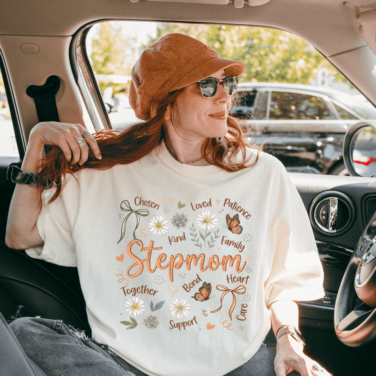 A woman wearing a white t-shirt with a design and text, sitting in a car with a hat on her head.