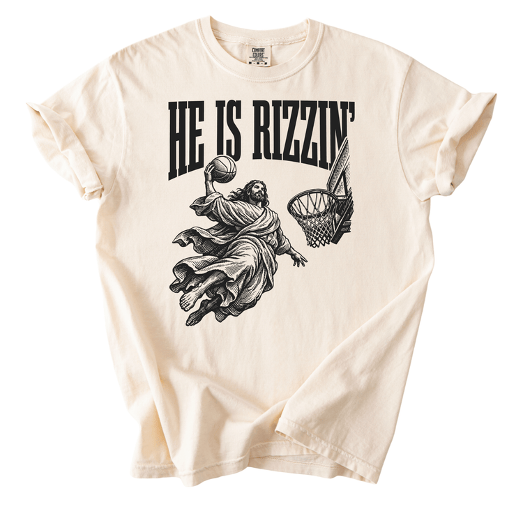 A white t-shirt with a graphic design depicting a person in a flowing robe holding a basketball, with the text "HE IS RIZZIN" above the image.