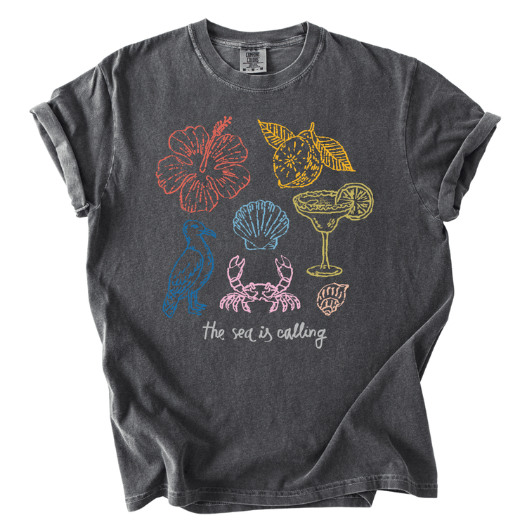 A gray t-shirt with a graphic design featuring various sea-themed illustrations, including a hibiscus flower, a cocktail glass, a seashell, a crab, and a bird. The text below the illustration reads "The sea is calling".