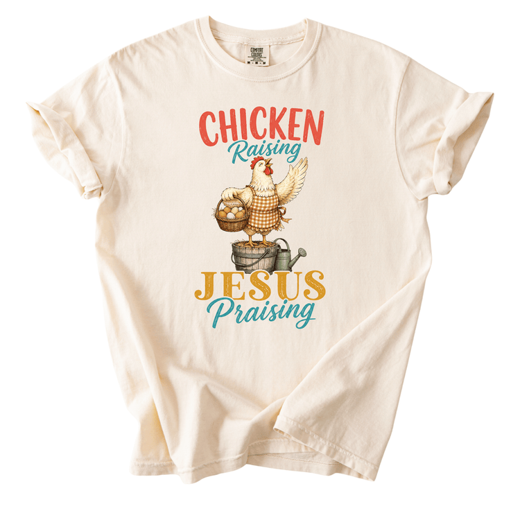 A white t-shirt with a graphic design depicting a chicken standing on a barrel and holding a basket of eggs, with the text "CHICKEN RAISING JESUS PRaying" printed on it.