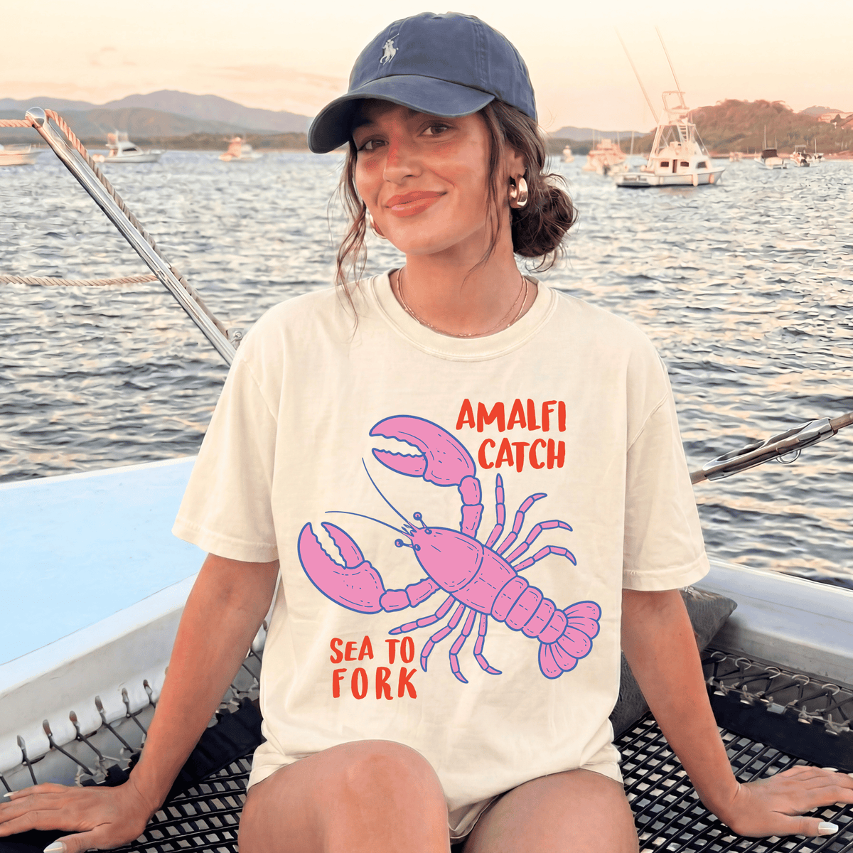 A woman wearing a white t-shirt with a pink lobster graphic and the text "Amalfi Catch Sea to Fork" is sitting on a boat in the water.