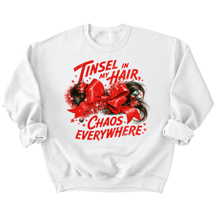 A white sweatshirt with a red graphic design featuring a red bow and the text "Tinsel in my hair, chaos everywhere" printed on it.
