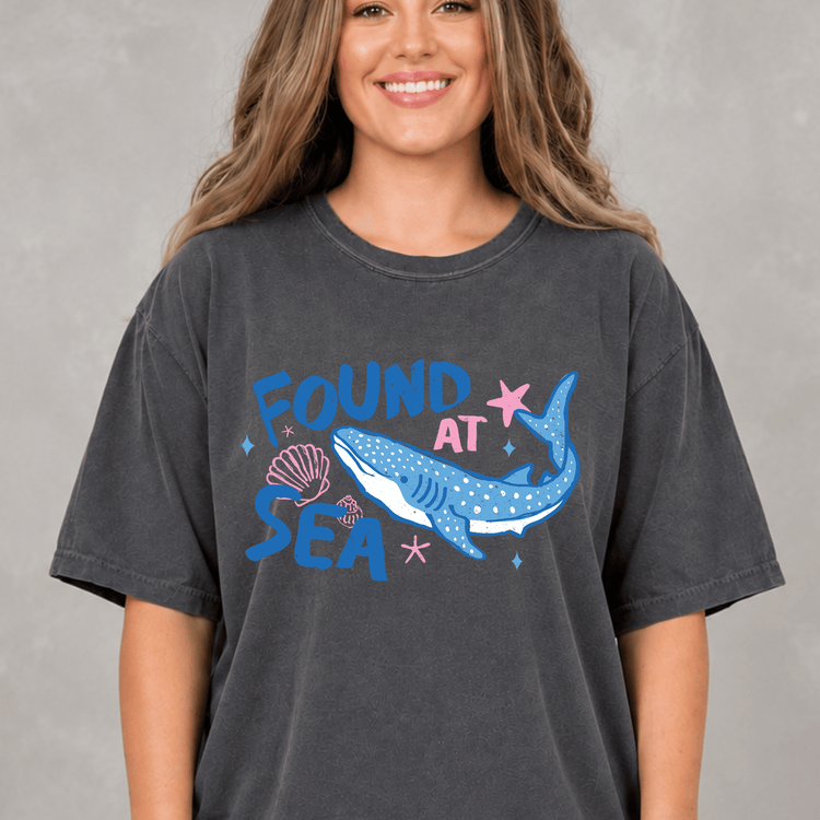 A woman wearing a gray t-shirt with a graphic design featuring a blue whale and the text "FOUND AT SEA" printed on it.