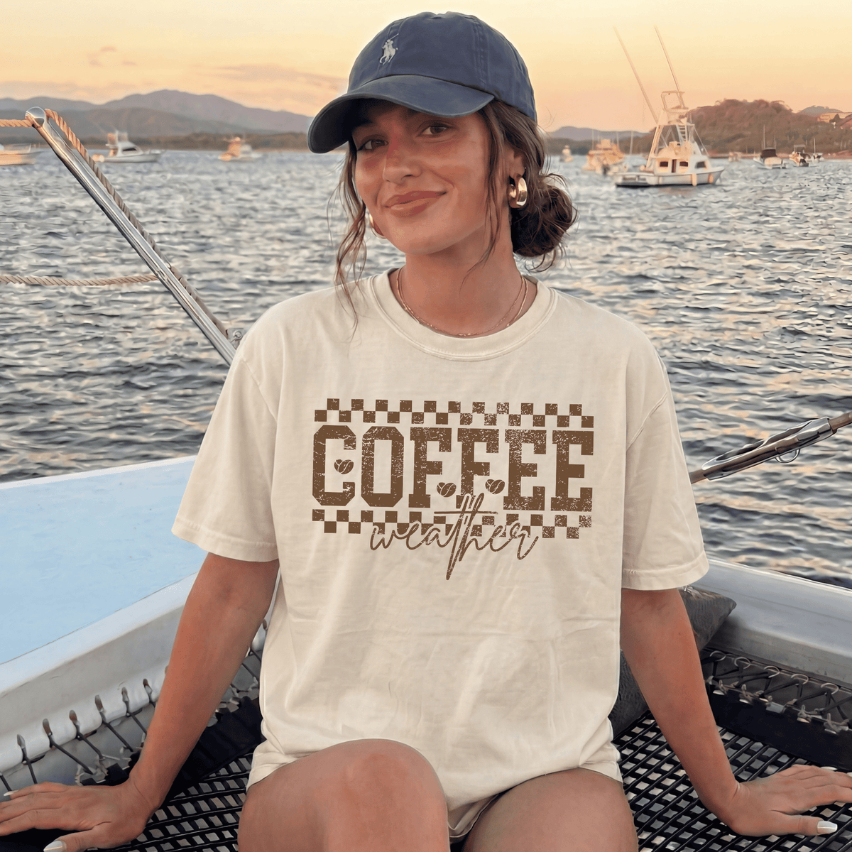 A woman wearing a baseball cap and a t-shirt with the text "COFFEE weather" is sitting on a boat, smiling at the camera.