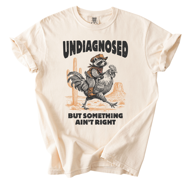 a t-shirt with a graphic design depicting a raccoon riding a rooster, with the text "UNDIAGNOSED BUT SOMETHING AIN'T RIGHT" printed on it.