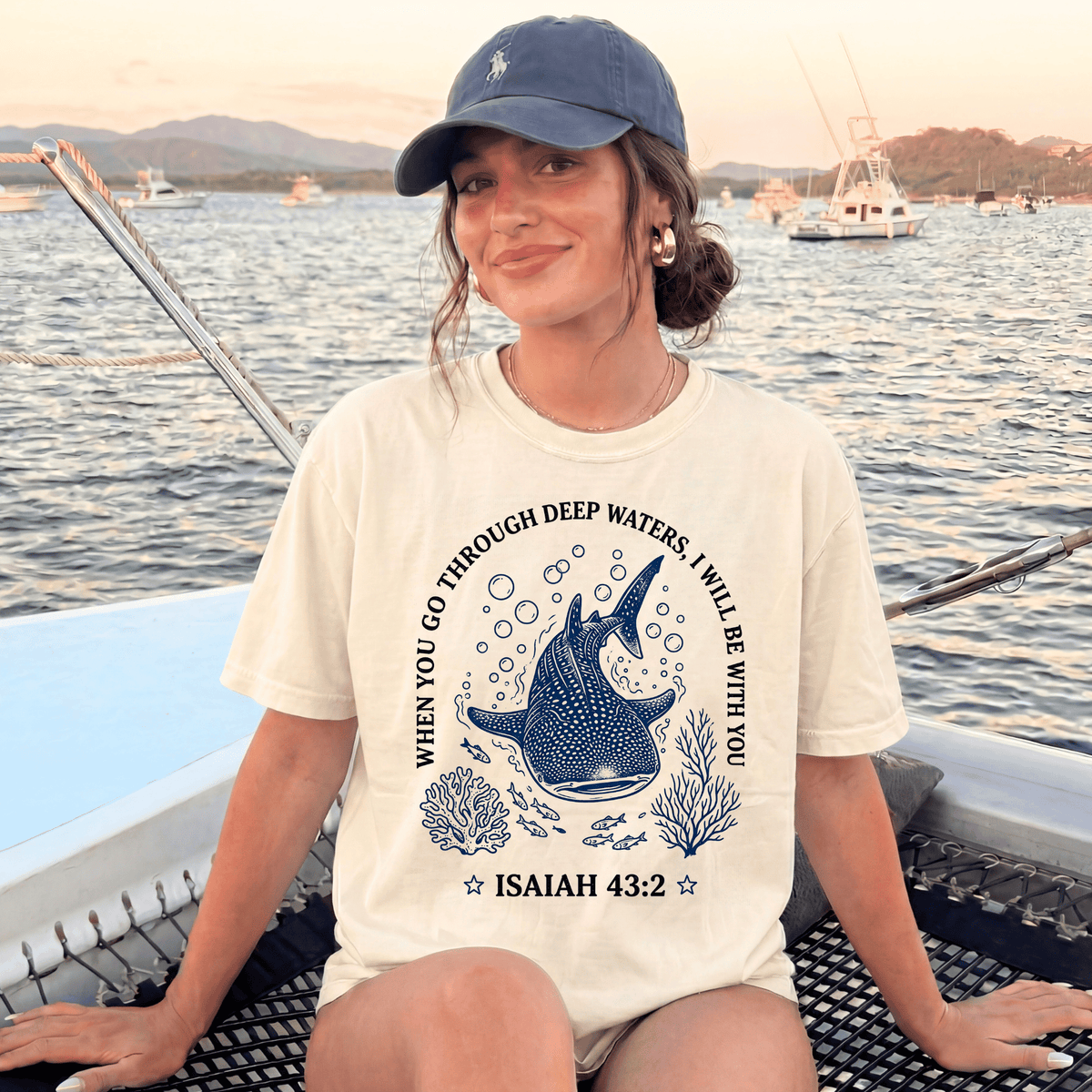 A woman wearing a white t-shirt with a graphic design of a blue whale and bubbles, sitting on a boat with a scenic background of mountains and boats.