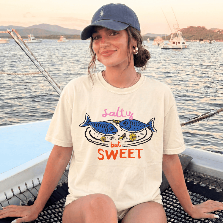 A woman wearing a white t-shirt with a graphic design and text, sitting on a boat in a body of water with other boats in the background.