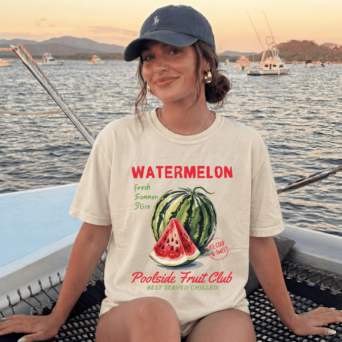 A woman wearing a white t-shirt with a watermelon graphic and text, sitting on a boat with a scenic background of mountains and boats.
