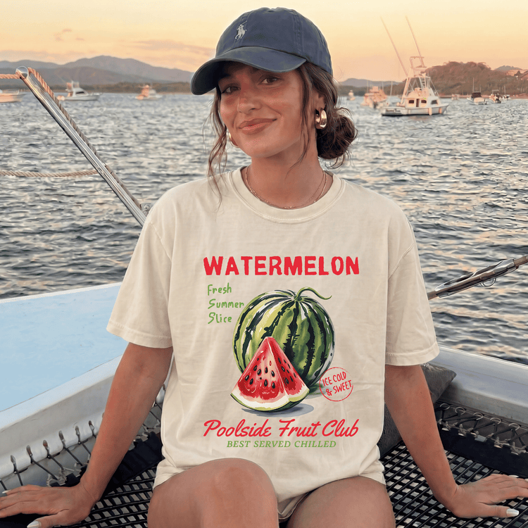 A woman wearing a white t-shirt with a watermelon graphic and text, sitting on a boat with a scenic background of mountains and boats.