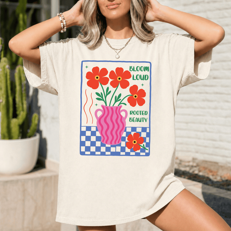 A woman wearing a white t-shirt with a graphic design featuring a pink vase with red flowers and the text "Bloom Loud Rooted Beauty".