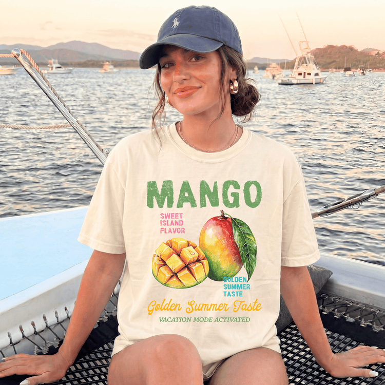 A woman wearing a cap and a t-shirt with a graphic of a mango and text promoting a "Mango" flavor, sitting on a boat with a scenic background of water and boats.