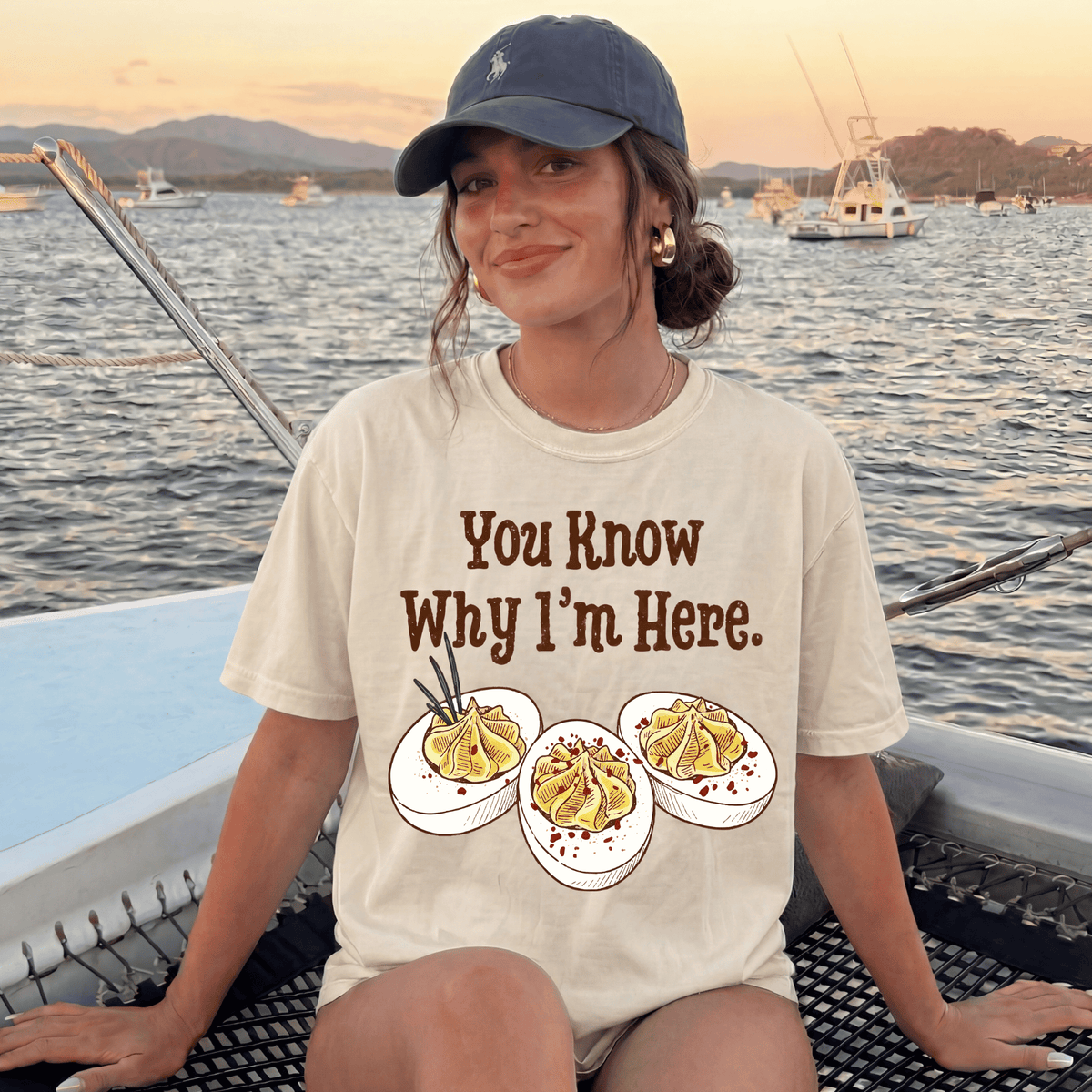 A woman wearing a white t-shirt with a graphic of three deviled eggs and the text "You Know Why I'm Here" is sitting on a boat in the water.