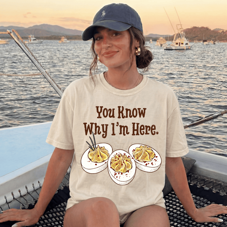 A woman wearing a white t-shirt with a graphic of three deviled eggs and the text "You Know Why I'm Here" is sitting on a boat in the water.