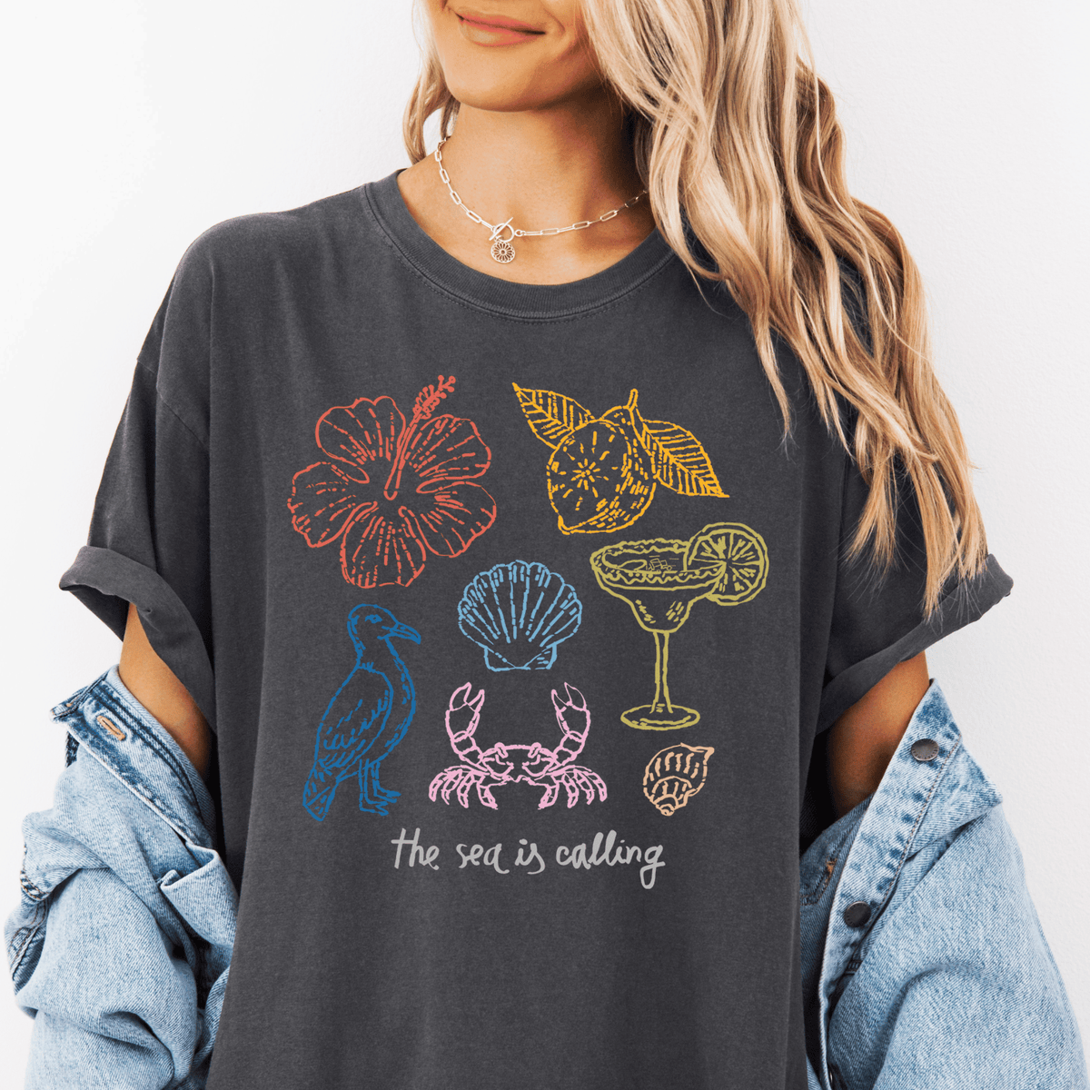 A woman wearing a gray t-shirt with a graphic design of various sea-themed illustrations, including a hibiscus flower, lemon, cocktail glass, and shell.