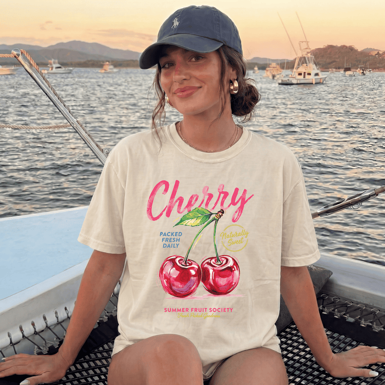 A woman wearing a white t-shirt with a graphic of cherries and text, sitting on a boat with a scenic background of water and boats.