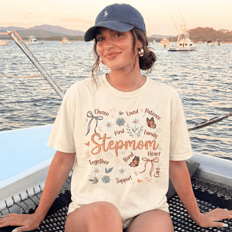 A woman wearing a white t-shirt with a graphic design and text, sitting on a boat with a scenic background of mountains and other boats.