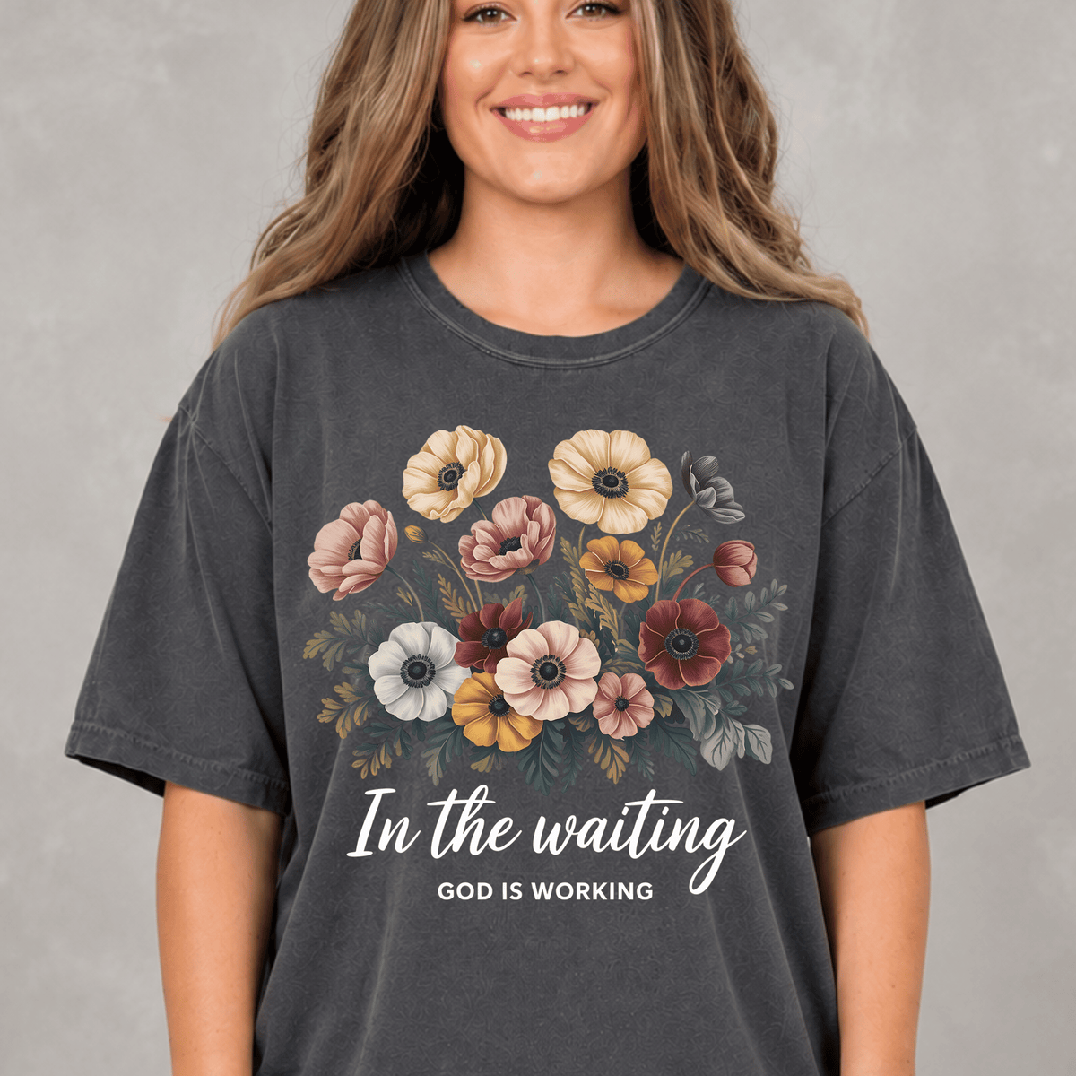 A woman wearing a gray t-shirt with a floral design and the text "In the waiting, God is working" printed on it.