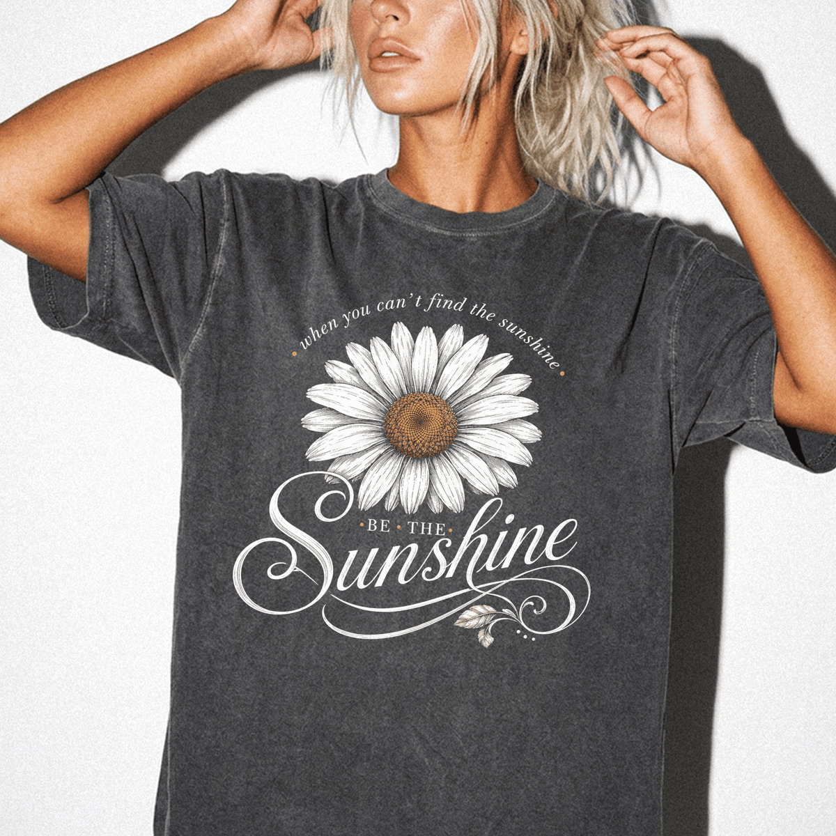 A woman wearing a gray t-shirt with a white daisy flower design and the text "Be the sunshine" printed on it.