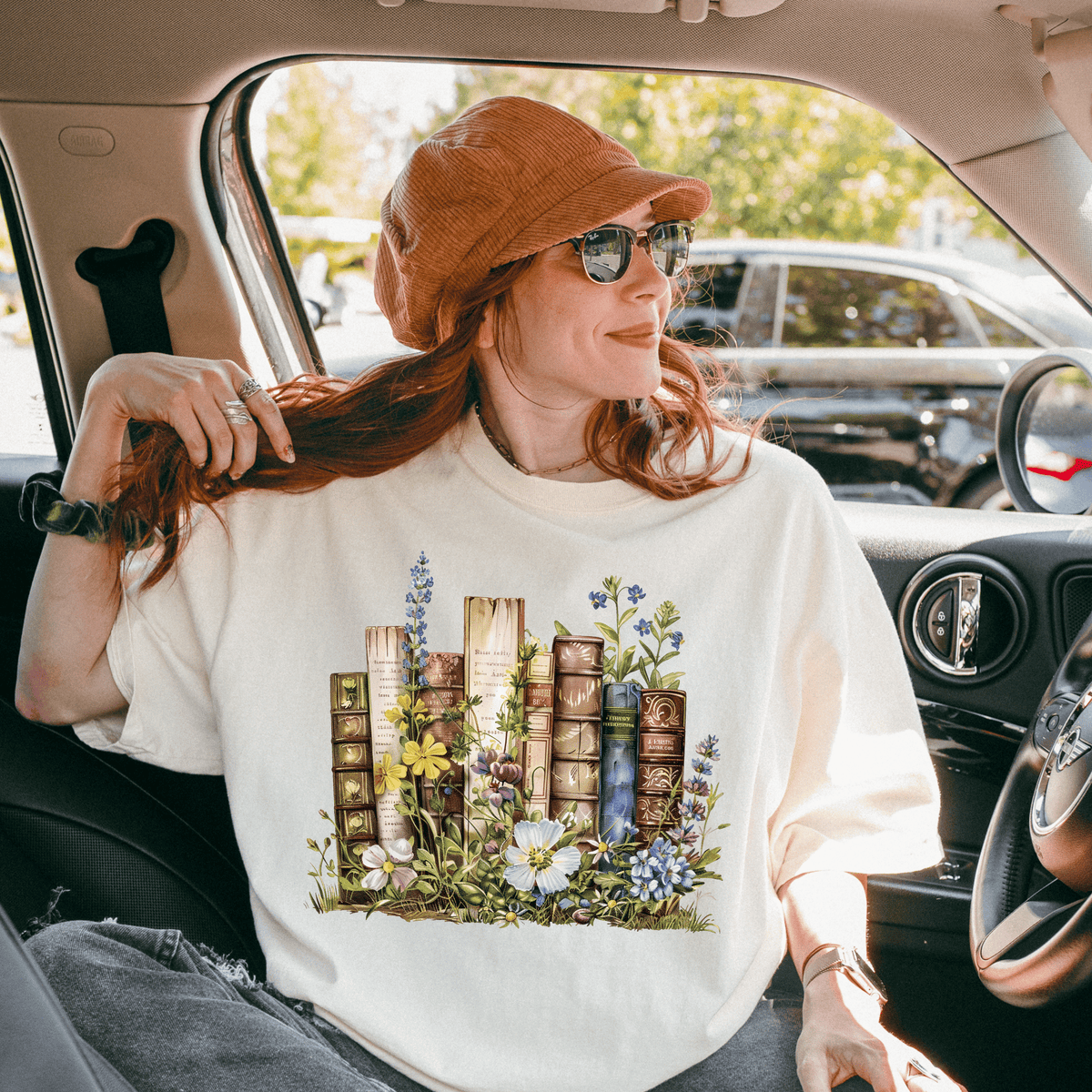 A woman with red hair is sitting in a car, wearing a white t-shirt with a floral design and a brown hat.