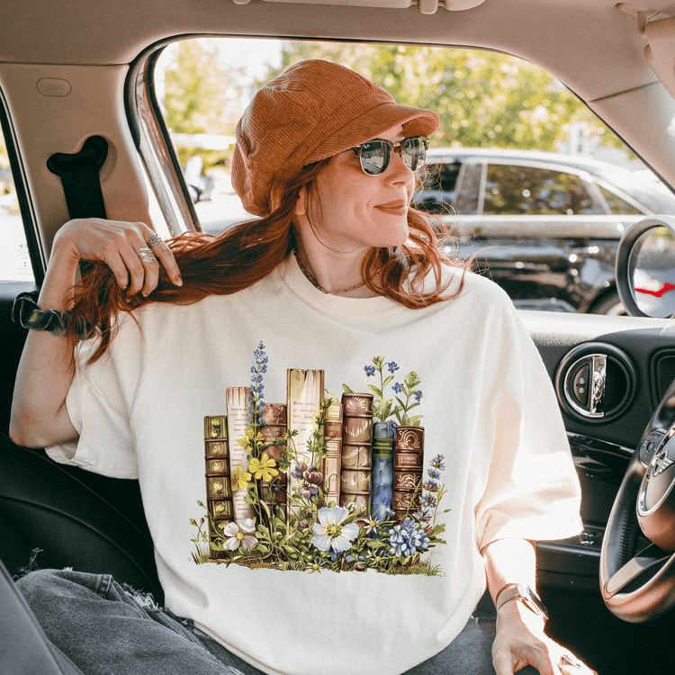 A woman with red hair is sitting in a car, wearing a white t-shirt with a floral design and a brown hat.