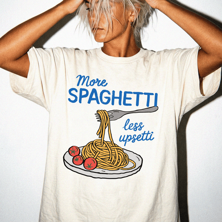 A person wearing a white t-shirt with a graphic design of spaghetti and tomatoes, along with the text "More Spaghetti less Upseti".