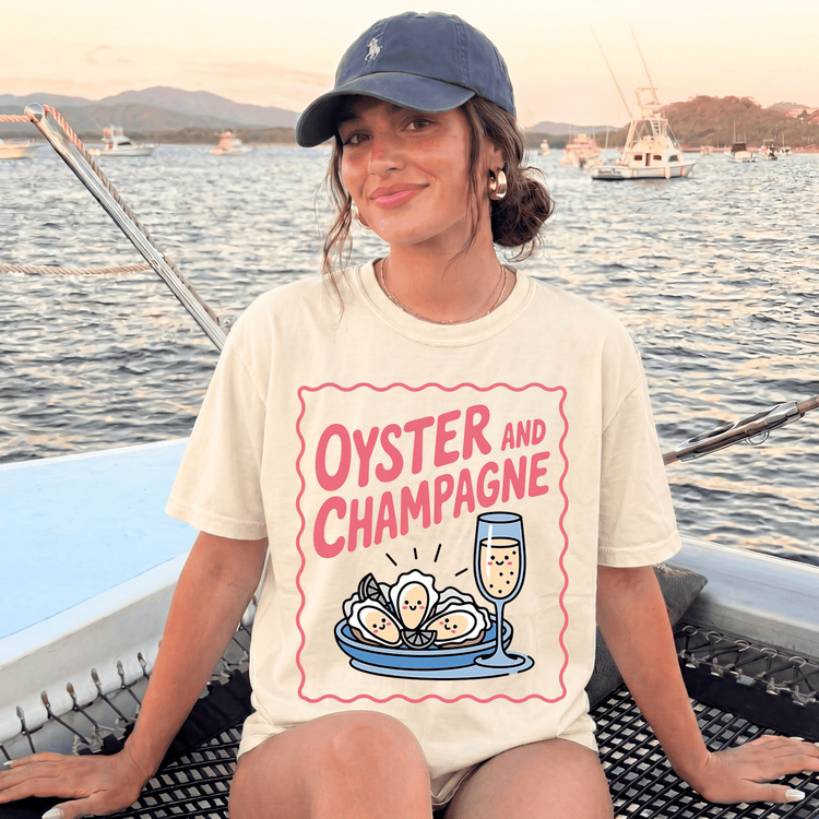 A woman wearing a hat and a white t-shirt with a graphic of oysters and champagne glasses, sitting on a boat in the water.