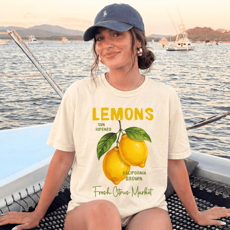 A woman wearing a cap and a white t-shirt with a graphic of lemons is sitting on a boat in the water.