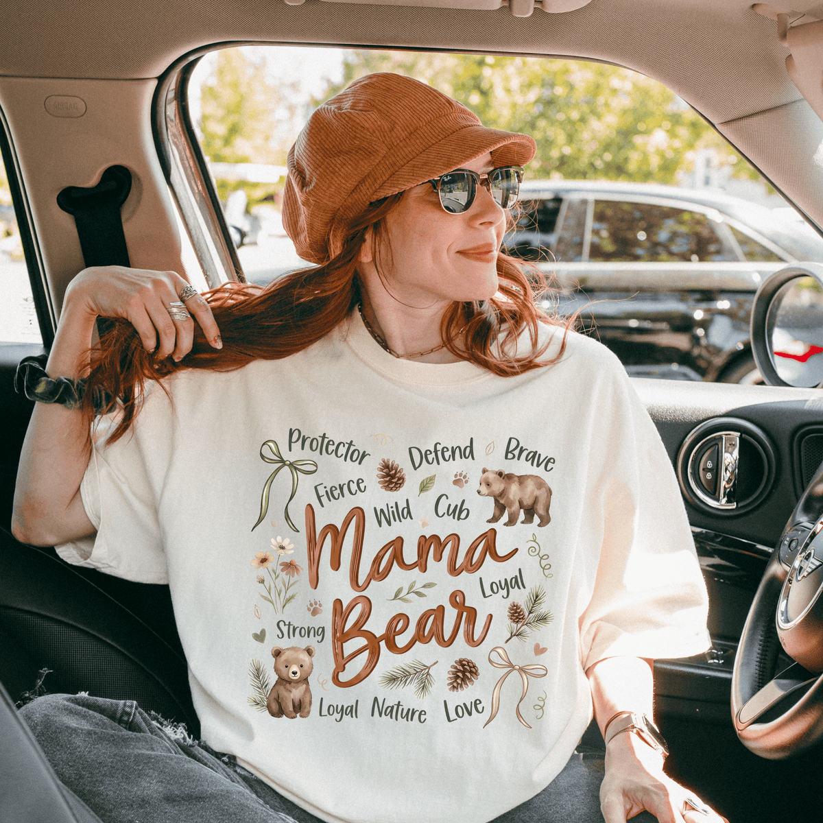 A woman wearing a white t-shirt with a design of a bear and the words "Mama Bear" is sitting in a car.