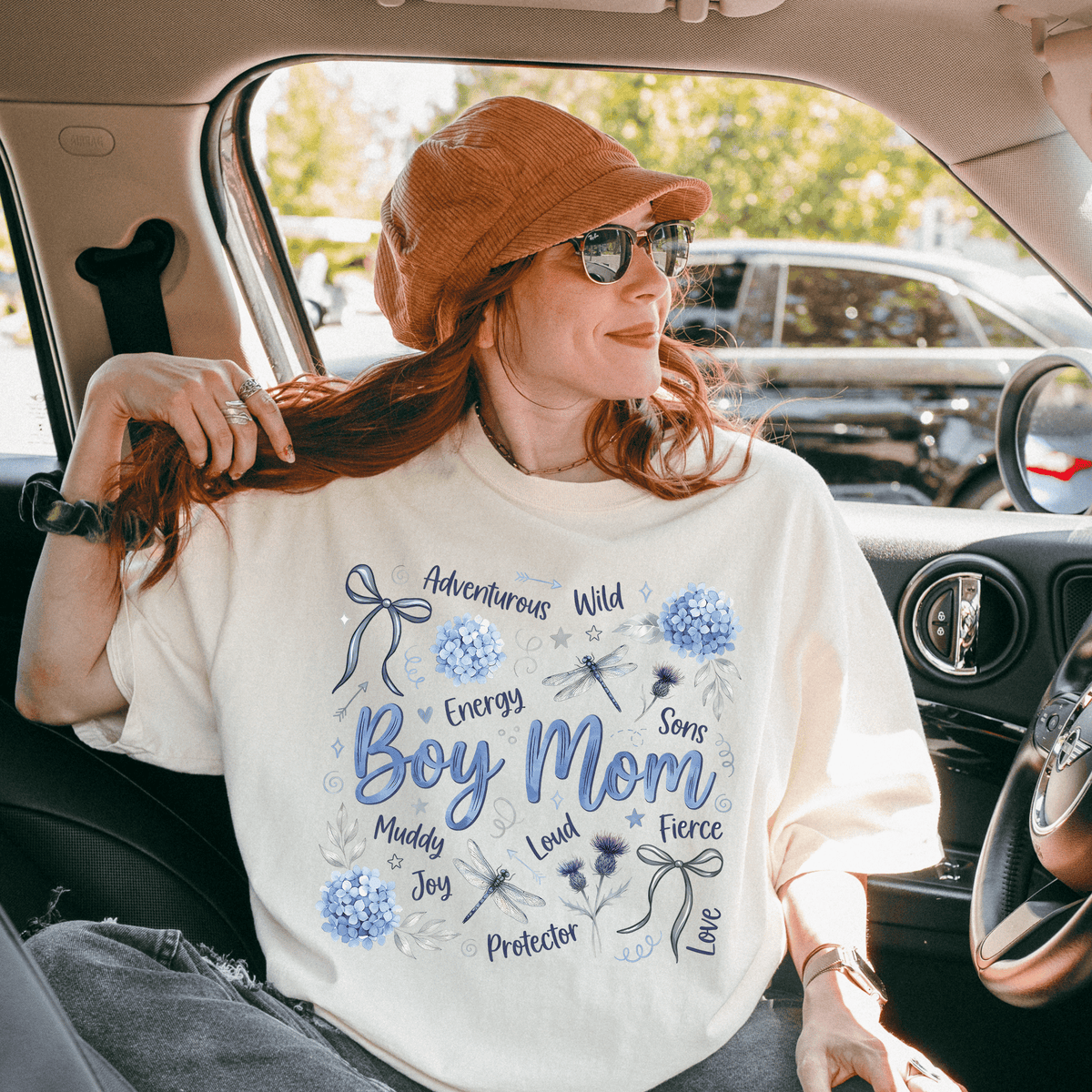 A woman wearing a white t-shirt with a graphic design and text, sitting in a car with a hat on her head.